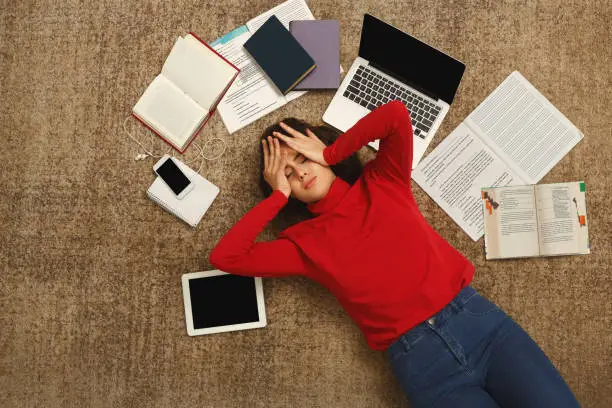 Tired student girl lying on the floor with books and gadgets Thesis Writing Mistakes